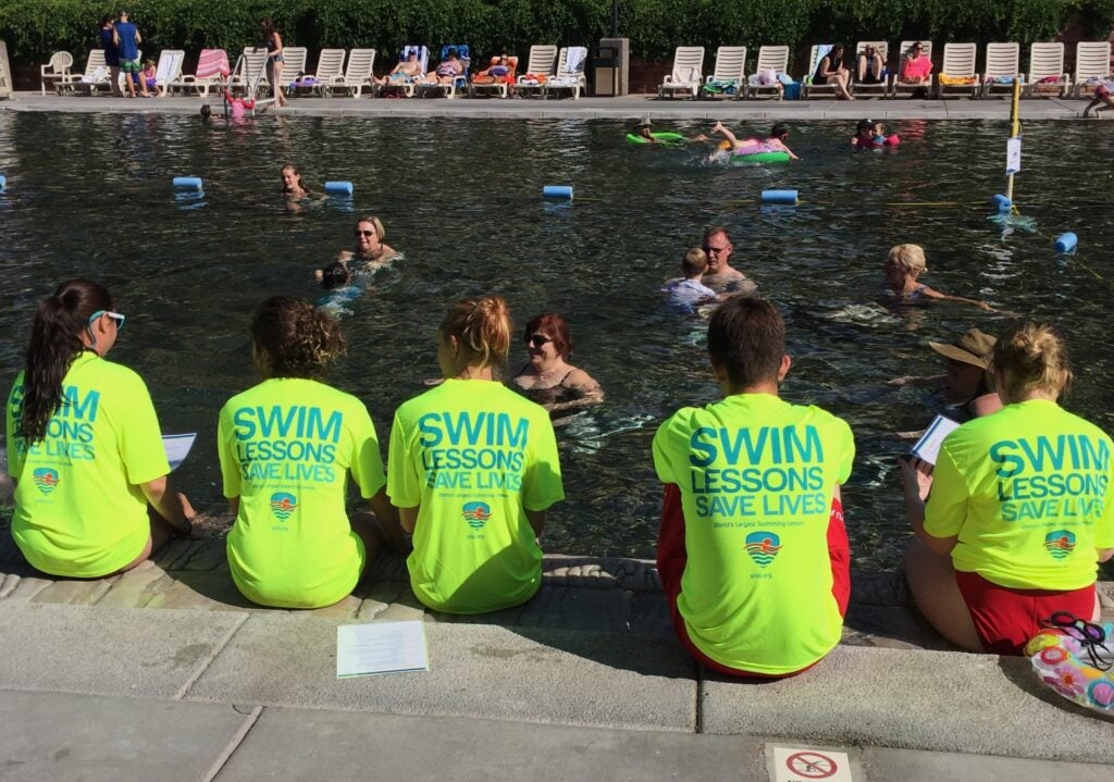 Swim instructors sit at edge of pool with t-shirts reading "Swim Lessons Save Lives"